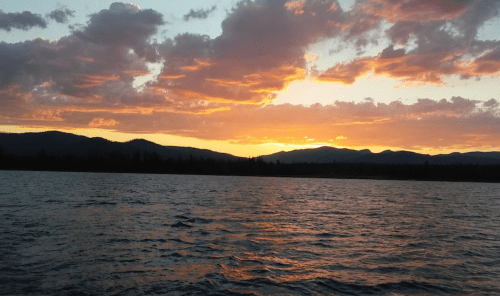 Sunset over a calm lake, with vibrant orange and pink clouds reflecting on the water and mountains in the background.