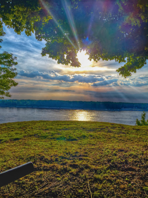A serene riverside view with sunlight streaming through clouds, illuminating the water and surrounding greenery.