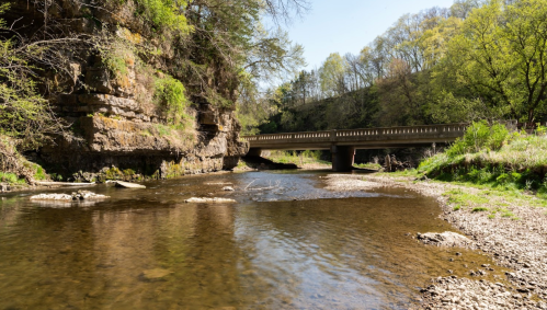 A serene river scene with a bridge, surrounded by lush greenery and rocky cliffs under a clear blue sky.