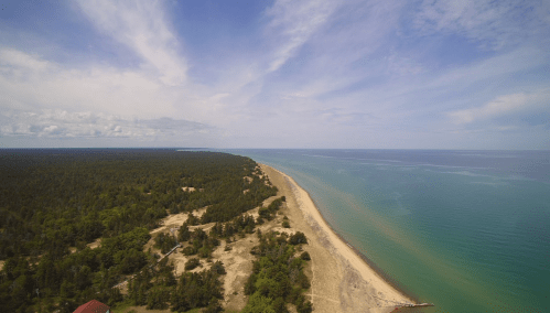 Aerial view of a sandy beach bordered by lush green trees and calm blue waters under a partly cloudy sky.