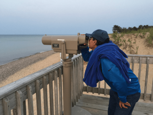 A person looking through a telescope on a beachside deck, with sand and water in the background.