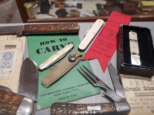 A collection of vintage knives, tools, and a green booklet titled "How to Carry," displayed with red ribbons and papers.