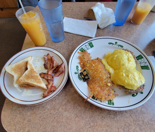 A plate with eggs Benedict, hash browns, toast, and bacon, alongside glasses of orange juice and water.