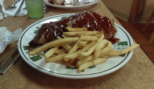 A plate of fries with a large piece of meat topped with ketchup, served in a casual dining setting.