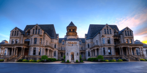 A grand, historic stone mansion with multiple towers and intricate architecture, set against a colorful sky at dusk.