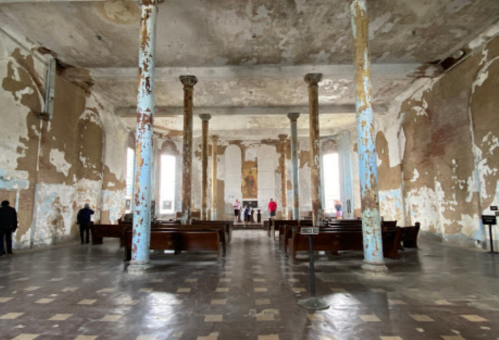 Interior of a dilapidated church with peeling walls, tall columns, and a few people gathered near the altar.