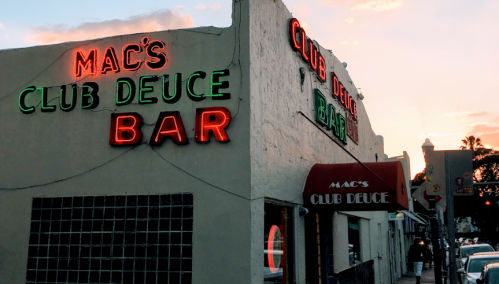 Exterior of Mac's Club Deuce Bar with neon signs at sunset, showcasing a lively atmosphere.