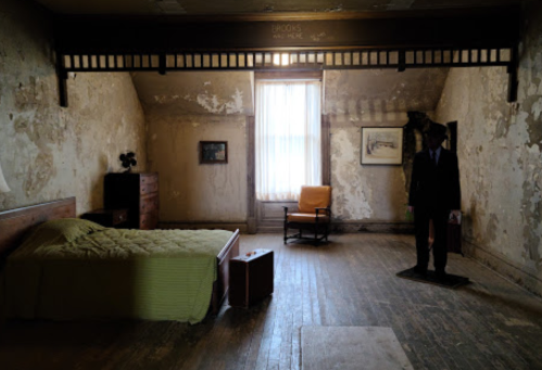 A dimly lit, worn bedroom with a bed, chair, and a figure in a suit standing in the corner. Peeling walls and wooden floor.