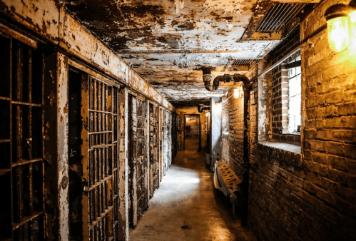 A dimly lit, rusty prison corridor with barred cells and exposed brick walls.