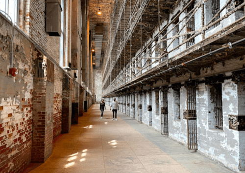 Two people walk down a long, narrow corridor of an old, abandoned prison with peeling walls and barred cells.