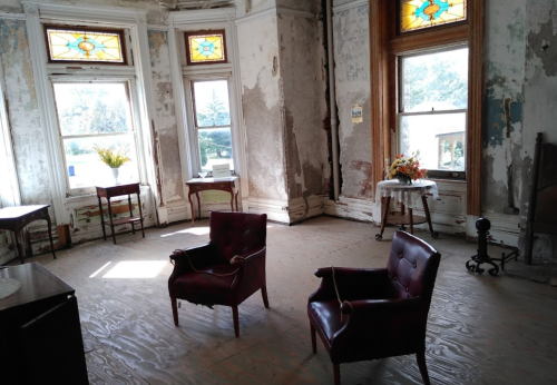 A sunlit, vintage room with two chairs, stained glass windows, and peeling paint on the walls.
