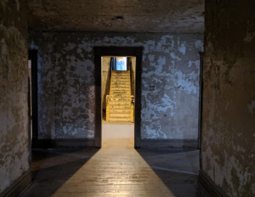 Dimly lit hallway with peeling walls, leading to a staircase in the background. Shadows create a mysterious atmosphere.