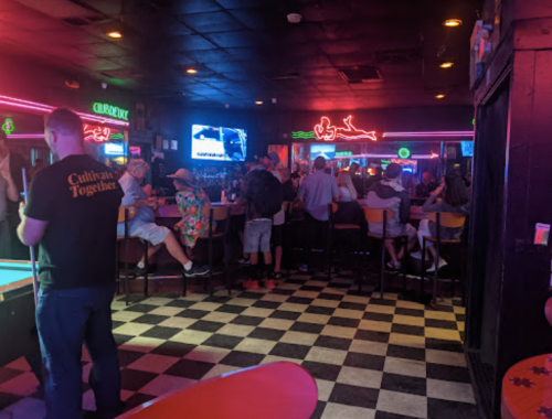 A lively bar scene with neon lights, patrons at the bar, and a pool table in the foreground.