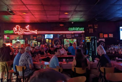 A lively bar scene with neon lights, patrons at the counter, and a vibrant atmosphere filled with conversation.