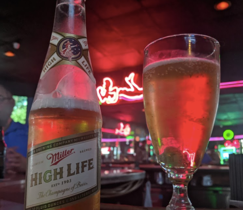 A bottle of Miller High Life beer next to a glass of beer, with a neon sign glowing in the background.