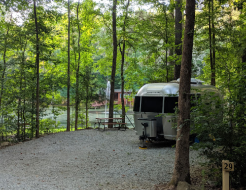 A silver Airstream trailer parked in a wooded area near a lake, with a picnic table and trees surrounding it.
