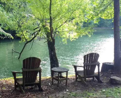 Two wooden chairs and a small table by a serene lake, surrounded by lush green trees.