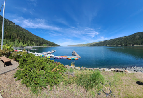A serene lake surrounded by mountains, with colorful kayaks lined up on a dock under a clear blue sky.