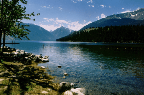 A serene lake surrounded by mountains, with clear water and rocky shores under a blue sky with scattered clouds.