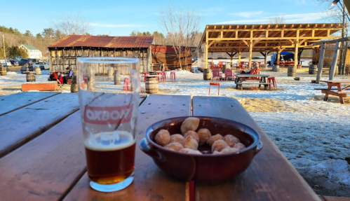 A glass of beer and a bowl of snacks on a wooden table, with an outdoor seating area in a snowy landscape.