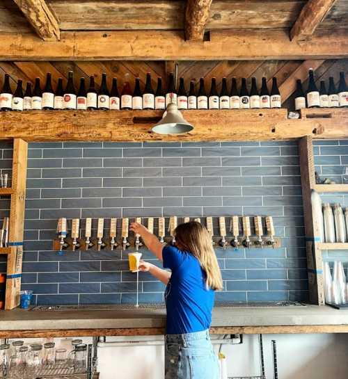 A woman in a blue shirt pours a beer from a tap at a rustic bar with bottles displayed above.