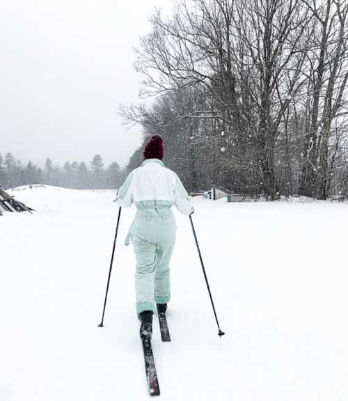 A person skiing on a snowy trail, wearing a mint green outfit and a red hat, surrounded by trees in a winter landscape.