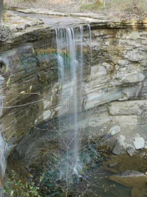 A serene waterfall cascading over rocky cliffs into a calm pool below, surrounded by trees and natural scenery.