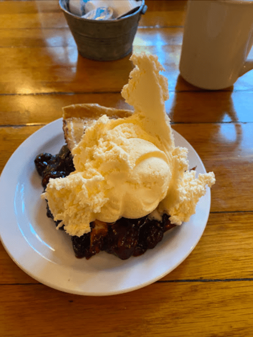 A slice of pie topped with a large scoop of vanilla ice cream on a white plate, set on a wooden table.