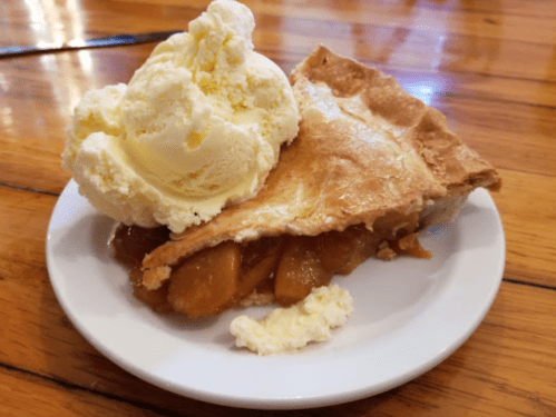 A slice of apple pie topped with a scoop of vanilla ice cream on a white plate, resting on a wooden table.