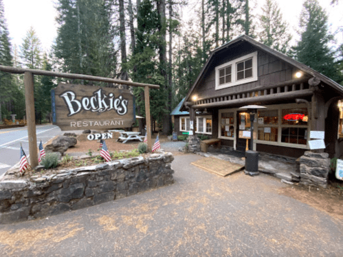 A rustic restaurant named Beckie's, surrounded by trees, with an "Open" sign and American flags at the entrance.