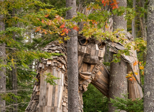 A large wooden sculpture resembling a giant creature stands among trees, surrounded by autumn foliage.