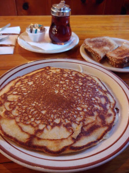 A large, golden pancake on a plate, with syrup and toast in the background on a wooden table.