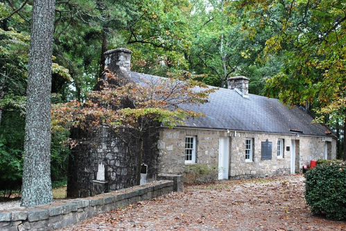 A rustic stone cottage surrounded by trees, with autumn leaves and a gravel path leading to the entrance.