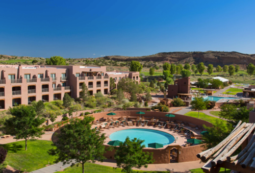 A scenic view of a resort with a circular pool, surrounded by green trees and mountains under a clear blue sky.