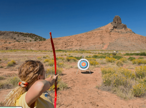 A child aims an arrow at a target in a desert landscape with a rocky formation in the background.