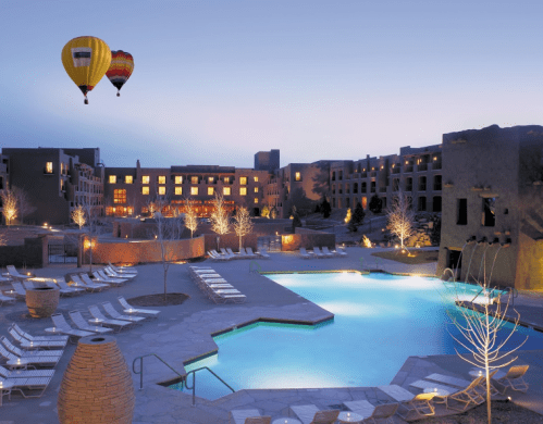 Hot air balloons float over a serene pool area at dusk, surrounded by lounge chairs and softly lit trees.
