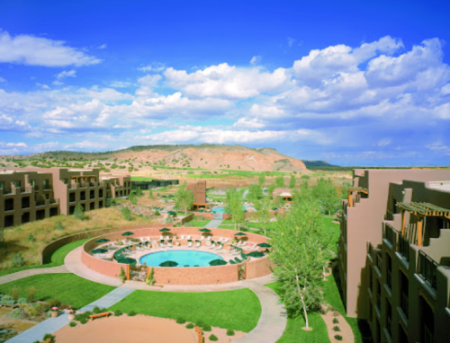 A scenic view of a resort with pools and landscaped gardens, set against a backdrop of hills and a blue sky.