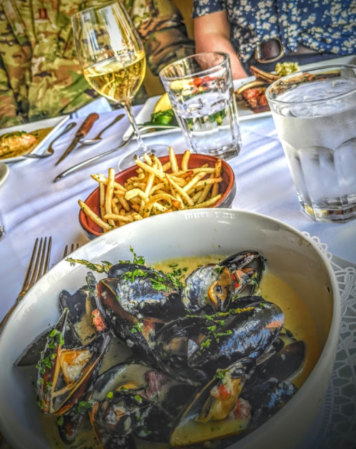 A bowl of mussels in a creamy sauce, accompanied by fries, glasses of water, and wine on a dining table.