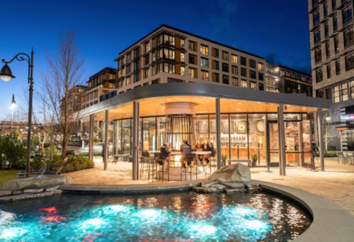 A modern building with outdoor seating by a pond, illuminated at dusk, surrounded by greenery and urban architecture.