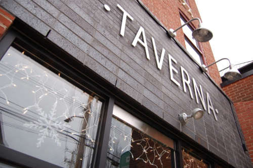 Sign for "Taverna" on a brick building, with decorative lights visible in the windows.