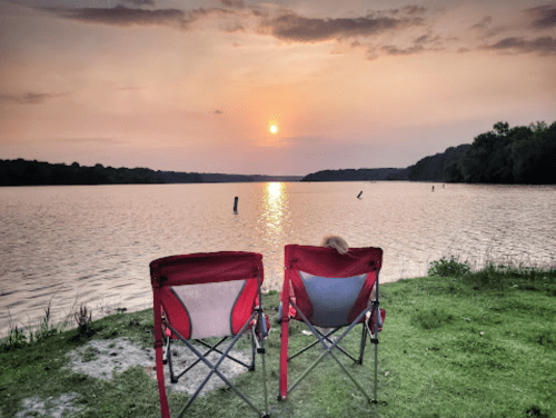 Two empty chairs facing a serene lake at sunset, with soft clouds and a peaceful atmosphere.