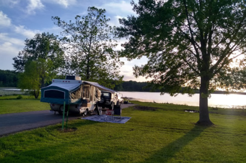 A camper parked by a lake, surrounded by trees, with a picnic setup on the grass under a clear sky.