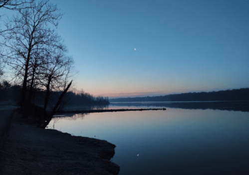 A serene river at dawn, with a crescent moon and trees silhouetted against a soft blue and pink sky.