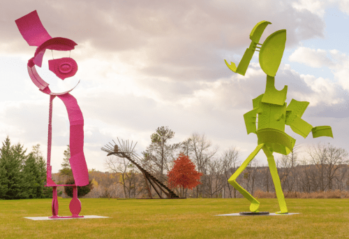 Two large, colorful metal sculptures stand in a grassy field under a cloudy sky, surrounded by trees.