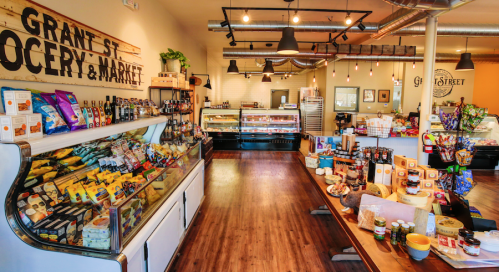 Interior of Grant St. Grocery & Market featuring shelves of gourmet foods, cheeses, and a display case. Warm, inviting atmosphere.