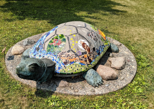 A large, colorful turtle sculpture adorned with various artistic designs, resting on a stone circle in a grassy area.