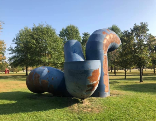 A large, abstract blue sculpture with curved shapes, set in a grassy park surrounded by trees.