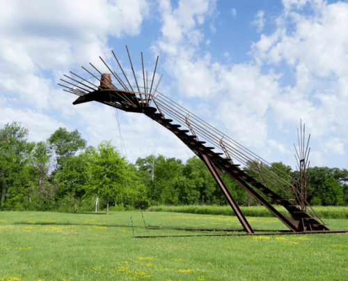 A large, abstract metal sculpture resembling a bird in flight, set against a grassy field and blue sky.