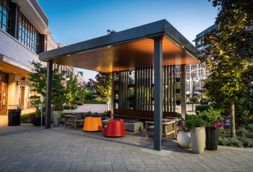 A modern outdoor seating area with colorful chairs, plants, and a wooden canopy, illuminated in the evening light.