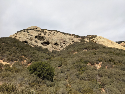 A rocky hillside with caves and sparse vegetation under a cloudy sky.